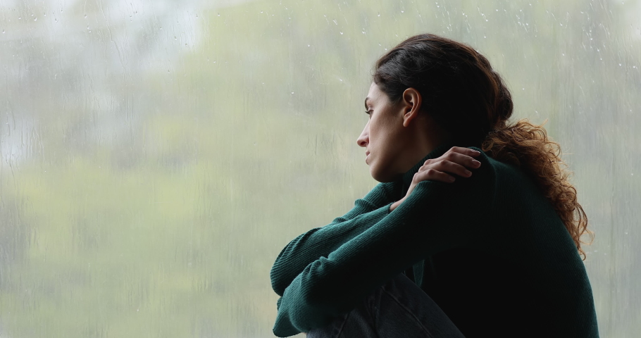 Sad melancholic young woman sitting on windowsill, cuddling knees, looking outside on gloomy rainy weather, suffering from loneliness or thinking of personal psychological problems or life troubles. | Shutterstock HD Video #1070487973