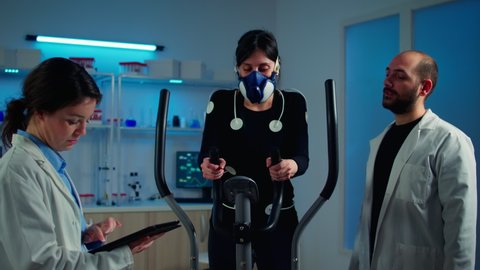 Woman athlete with mask doing fitness exercises in science sport lab with electrodes attached while scientist holding tablet supervising whole process. Physician using notepad controling EKG data