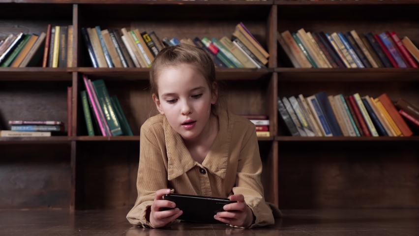 Interested schoolgirl plays online game on black smartphone sitting at brown table against large bookcase staying home at pandemic isolation