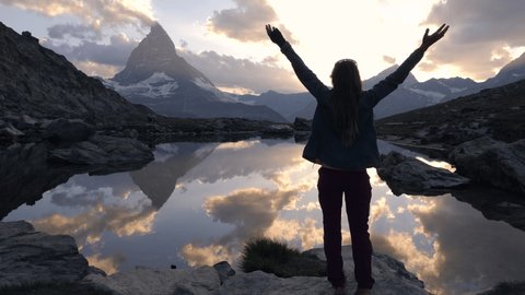 Slow motion: Young woman on a hike takes a moment to enjoy breathtaking views of mountains and lake at sunrise. Woman standing arms wide open in nature 