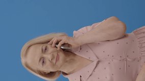 An elderly woman in front of a blue background speaks by mobile phone. Portrait of a fun elderly woman laughing with laughter talking on the phone.Video for the vertical story.