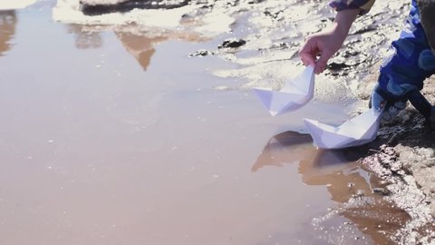 Children launches a paper boats into a muddy spring puddle.Spring concept.Children's leisure.Close-up, slow motion.