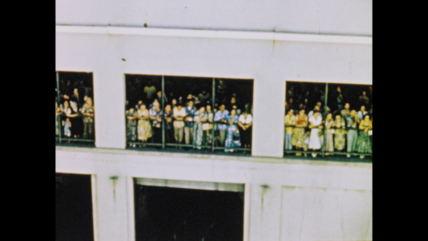 1950s: Girl in leis waves from deck of boat. People wait on dock. Aloha Tower with bell ringing. Honolulu street.