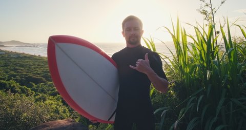 Surfer with board. Male caucasian surfer in black wetsuit stands on the grass holds the surf board and shows the shaka sign
