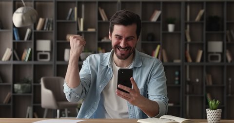 Overjoyed young handsome caucasian man feeling excited od getting mobile message with amazing news, making yes gesture celebrating online lottery giveaway win or successful auction betting indoors.