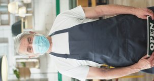 Portrait of senior grey-haired Caucasian male employee in apron wearing medical mask standing in coffee shop holding in hands Open sign, reopening in quarantine, business. Vertical video concept