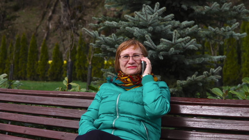 Cheerful senior woman having a phone talk sitting on the bench in the park on autumn or spring day