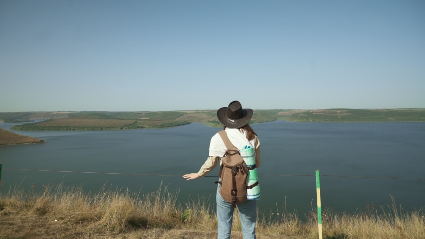 Back view of happy tourist in cowboy hat with backpack standing on edge of rock and enjoying beautiful view of Dniester river. National park in Ukraine.