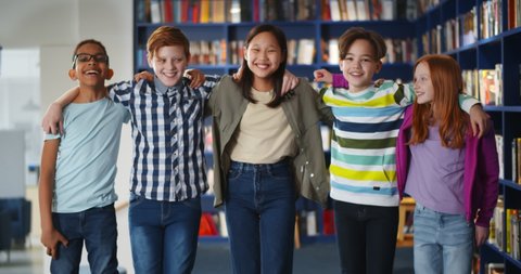 Portrait of multiethnic classmates hugging and smiling at camera in school library. Happy diverse pupils standing in row and embracing in library. Education and friendship portrait