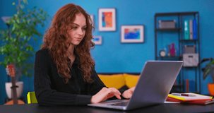 Beautiful Caucasian Girl with Curly Hair Sitting at a Desk in a Cozy Living Room and Using Laptop Computer at Home. She's Browsing the Internet and Checking Videos on Social Networks.