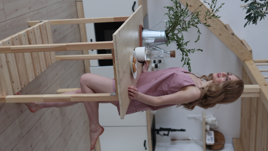 a woman in pajamas, top and shorts, leans on a wooden table in the kitchen. Vertical Video