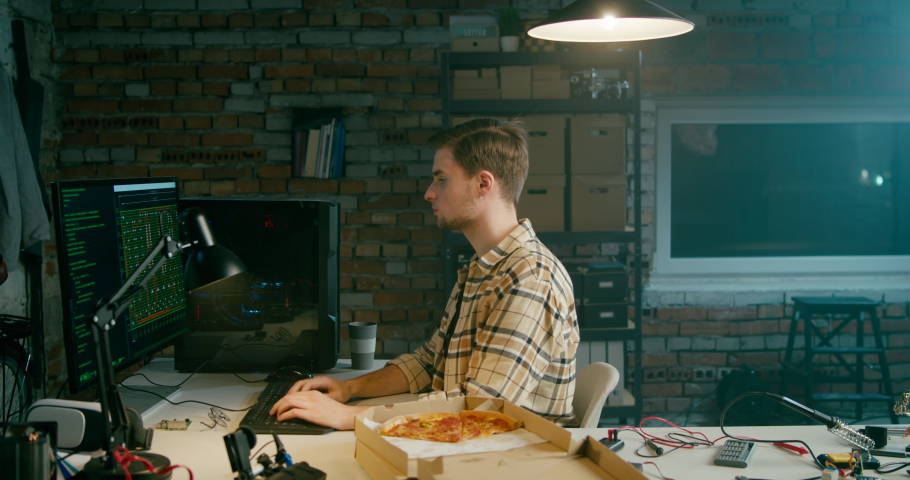 Coder Man or Hacker Eats Pizza Working on Computer in Loft Electronics Workshop or Home Garage at Night or Late Evening. Fast Food Lunch in IT Startup Office. 4K Wide Left Pan Shot Royalty-Free Stock Footage #1070527075