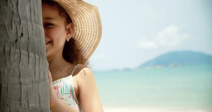 Portrait of a funny little girl in a hat, a smiling child looking at the camera, stands on the beach by the sea, a cute child with a beautiful face stands on the beach. Happy childhood concept.