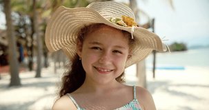 Portrait of a funny little girl in a hat, a smiling child looking at the camera, stands on the beach by the sea, a cute child with a beautiful face stands on the beach. Happy childhood concept.