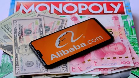 Alibaba logo seen on smartphone which is placed on money - US dollars and Chinese yuan, and Monopoly game on the blurred background. Concept. Staffrod, United Kingdom, April 10, 2021.