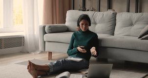 Full length relaxed young pleasant woman sitting on floor near sofa, wearing headphones, communicating distantly with mentor teacher using video call application on computer, preparing for exams.