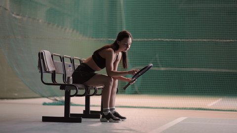 sportswoman is wearing sportswear is sitting on bench on tennis court after match or training, sporty person