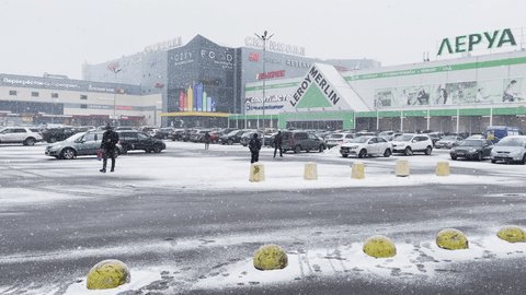 Russia, St.Petersburg, 18 March 2021: Heavy snowfall in city bustle, an automobile parking at huge shopping center, a snow storm, blizzard, multi-colored signs on the building