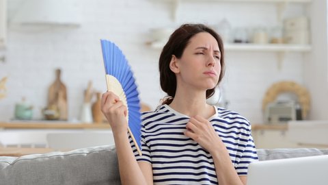 Tired millennial woman suffers from stuffiness and an inoperative air conditioner, waving blue fan sitting on couch at home working on laptop computer. Overheating high temperature, hot summer weather