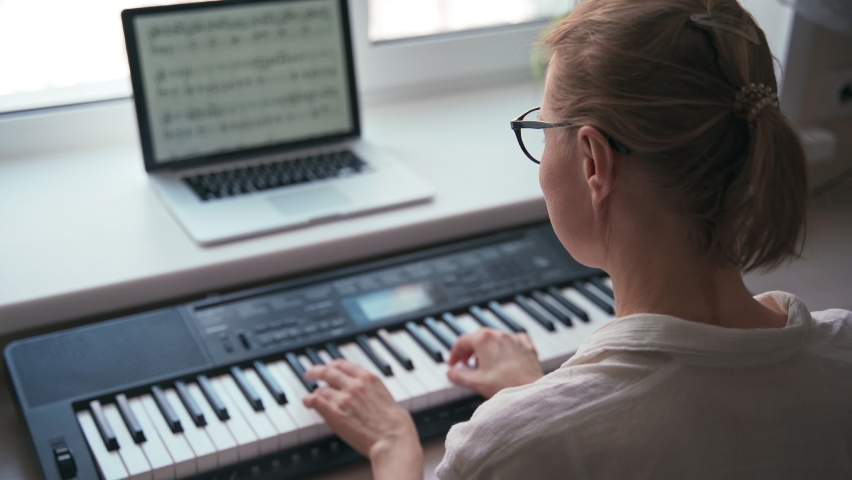 Back shot of a young woman playing on a piano keyboard and reading sheet music from a laptop screen.