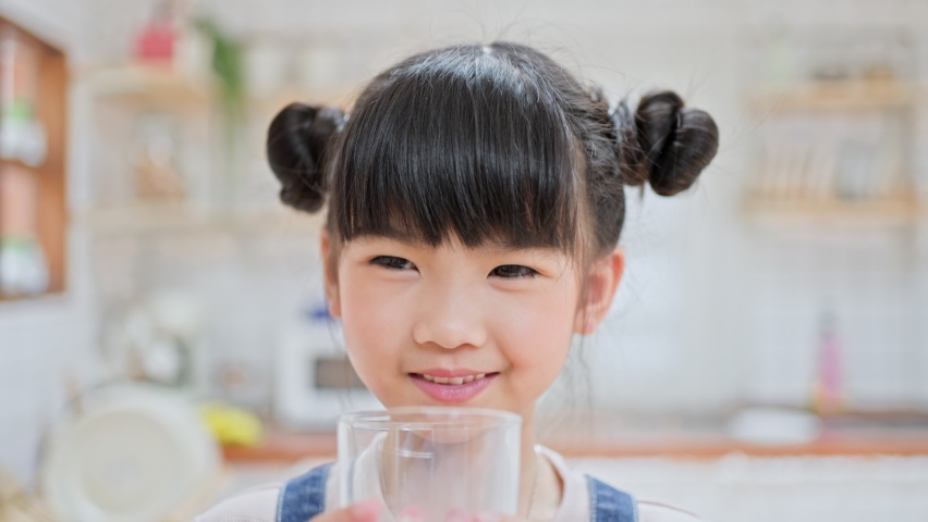 Portrait of Asian little cute kid holding a cup of milk and drinking in kitchen at home. Young preschool girl child or daughter messing up milky on moustache and lips then looking at camera in house. Royalty-Free Stock Footage #1070541238