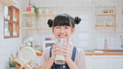 Portrait of Asian little cute kid holding a cup of milk and drinking in kitchen at home. Young preschool girl child or daughter messing up milky on moustache and lips then looking at camera in house.