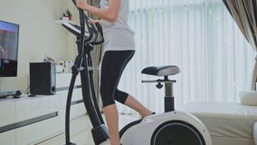 Asian Sport woman workout exercising by watching fitness live or video tutorial TV online on television at home. Casual young Girl using exercise bike follow instruction from trainer during quarantine