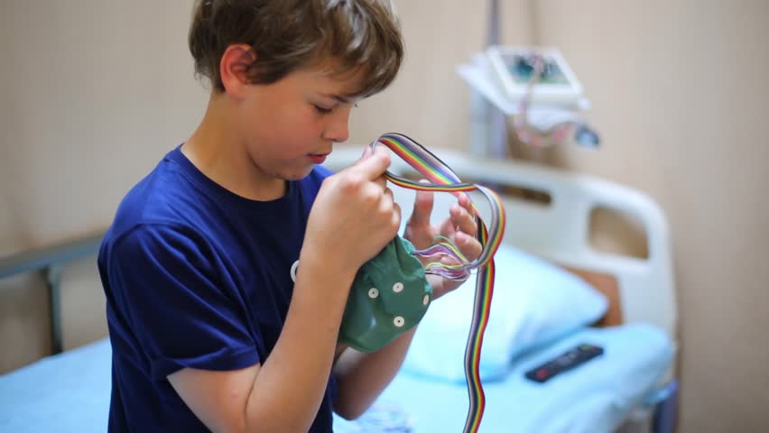 Boy sits on bed and looks at special cap for electroencephalography he holds in hands.