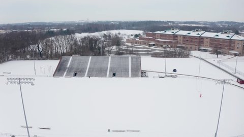 Aerial of school athletic fields and bleachers covered in winter snow. University college campus in United States of America during snowstorm. Aerial drone overhead view.