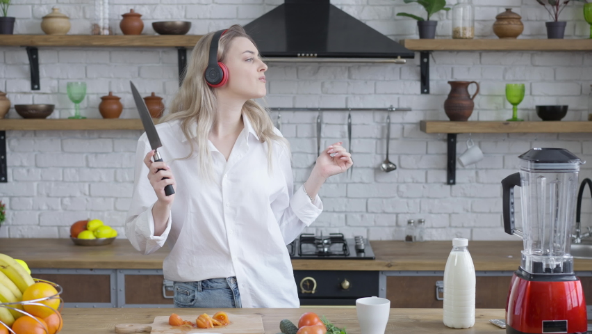 Portrait of cheerful beautiful young woman in headphones dancing and singing as cooking healthful vegan salad in kitchen at home. Joyful happy Caucasian millennial having fun indoors. Vegetarianism