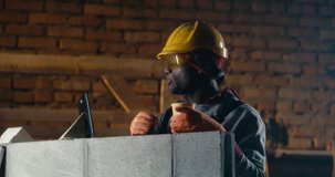 Black man in protective uniform enjoying hot drink and cracker and making video call to friend against brick wall during lunch break on construction site