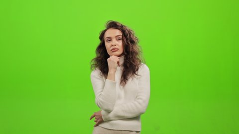 The woman is standing and looking at the camera. She is thinking and holding her hand to her chin. She is standing on a green background. Green screen. 4K