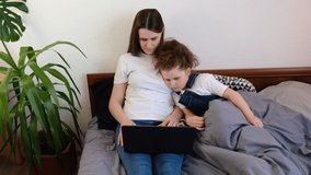 Young mother with little kid daughter watching video cartoons or playing game on laptop together, babysitter or nanny teaching child girl to use computer sit on bed at home. Device overuse concept