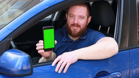 European bearded man in blue shirt sitting in the blue car, smiling and showing a mobile phone with green screen