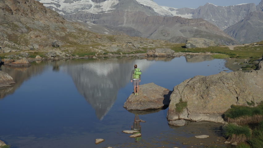 Young hiker stands on a rock in the middle of an Alpine lake harms wide open. Man contemplating nature 