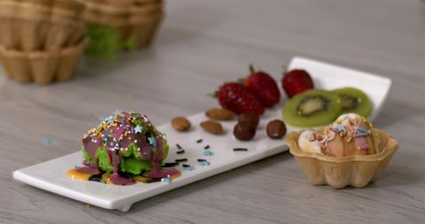 Chocolate pods fall on popsicles on a plate, multicolored and multi-flavored ice cream, kiwi, lemon, strawberry. Cold dessert on a wooden table. Slow motion, filmed high speed cinema camera. 4K.