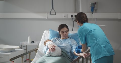 Young mother holding newborn baby while nurse set catheter in her hand. Woman after childbirth resting in hospital bed with infant baby and medical worker adjusting iv drip for her