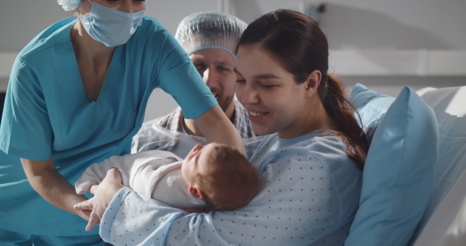 Couple holding newborn baby in maternity hospital after childbirth. Cropped shot of nurse giving infant baby to happy mother lying in hospital be and father sitting near | Shutterstock HD Video #1070576323