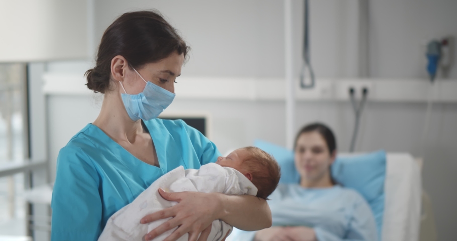 Portrait of nurse wearing safety mask cradling newborn in arms with young mother resting in bed at hospital ward. Happy woman relaxing in bed after childbirth with doctor holding baby Royalty-Free Stock Footage #1070576365