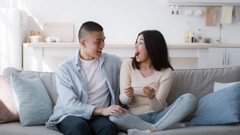 Joyful Asian Couple Holding Positive Pregnancy Test Hugging And Celebrating Result Sitting On Couch At Home. Young Japanese Family Awaiting Baby. Childbirth And Happy Family Moments Concept