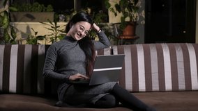 Happy attractive woman sitting on couch in cozy living room in evening with laptop video chat with friends. Asian girl brunette laughing resting with computer at home on couch writing in chat messages