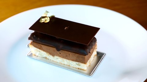 Chef Woman Hand Place a Hazelnut on Opera Cake Desserts on White Plate in Restaurant. Decoration of Chocolate Piece of Cake with Edible Gold and Nut in Cafe