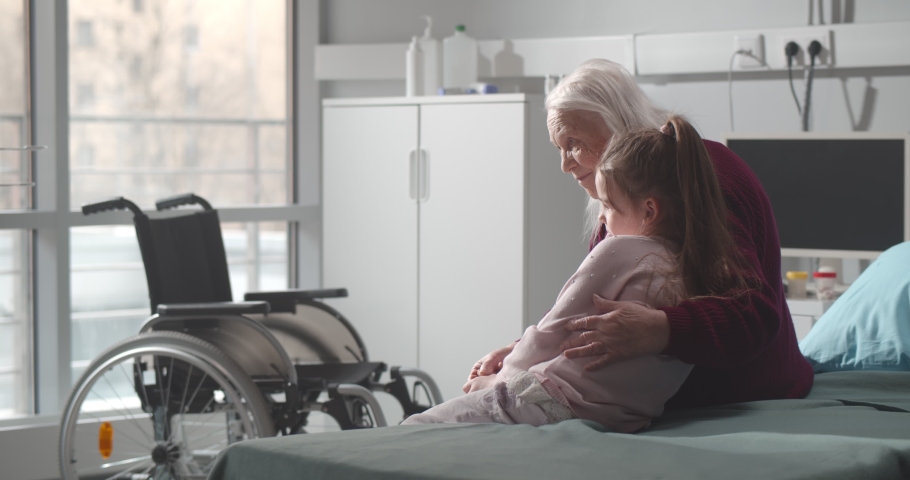 Senior woman and little girl sitting on hospital bed and hugging. Side view of grandmother and granddaughter embracing resting in nursing home or hospital ward