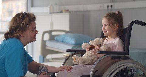 Child patient in wheelchair sitting in hospital ward talking to female nurse. Medical worker entertaining and cheering little disabled girl in wheelchair