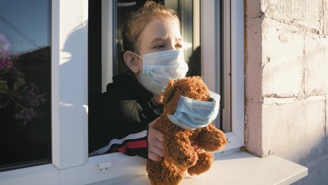 Quarantine, threat of coronavirus. Sad child and his teddy bear. Bored girl wears medical mask in home quarantine coronavirus looks out of the window. Epidemic covid 19 prevention concept.