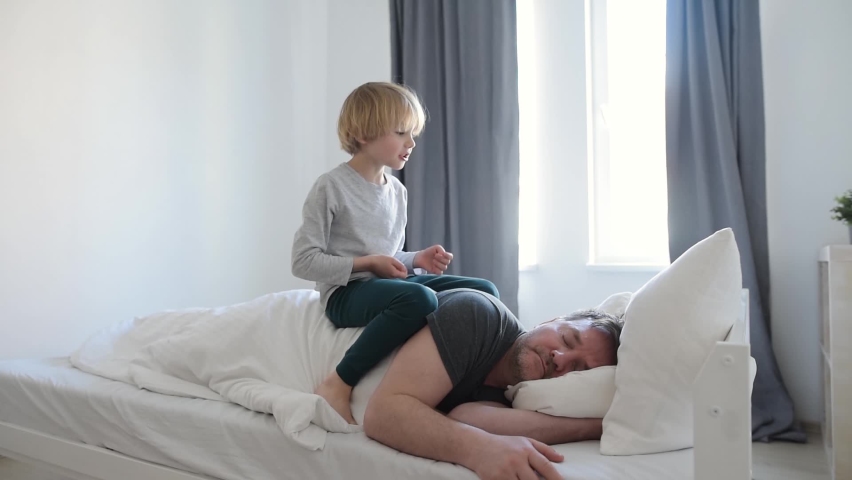 Exhausted parent with hyperactive child. Chaos when kids at home during long weekend or lockdown. Little boy try wakes his tired father. Dad sleep early morning in sunny bedroom during his son awake Royalty-Free Stock Footage #1070598988
