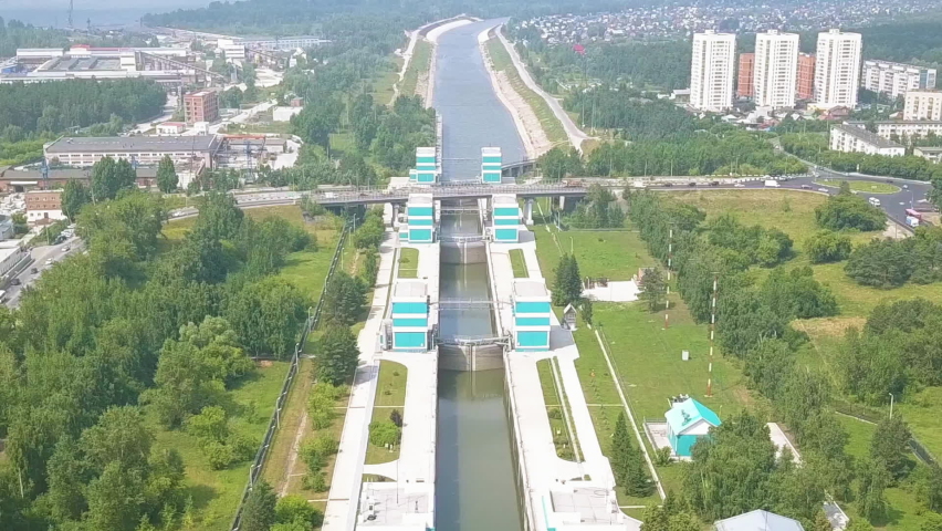 The shipping gateway of the Novosibirsk hydroelectric power station on the Ob River is captured in an aerial view, revealing a multi-chamber lock system flanked by turquoise and white control towers. The structure spans a wide canal that cuts through a lush green landscape, with a road bridge crossing overhead and residential high-rises visible in the distance. The gateway is divided into three chambers to allow vessels to pass the dam, which forms the Novosibirsk Reservoir, the largest artificial lake in Siberia at the time of its construction in 1956. The surrounding area is dense with summer foliage, and the water in the canal is calm. The camera slowly pulls back while zooming in, creating a subtle shift in perspective that emphasizes the scale of the engineering installation against the urban and natural backdrop. The scene is set in bright daylight under clear skies, conveying a sense of industrial functionality integrated into a verdant riverside environment. Dolly zoom.