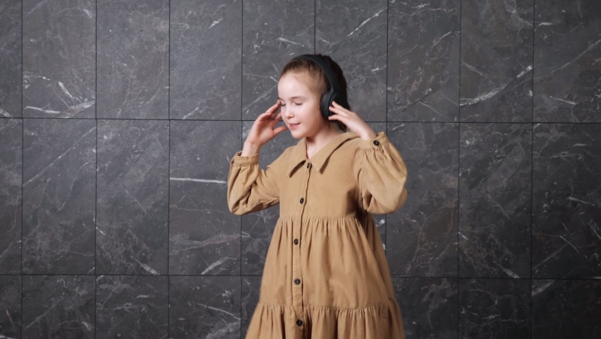 Excited schoolgirl listens to music in large black headphones and dances with delight against decorative grey white marble wall at school