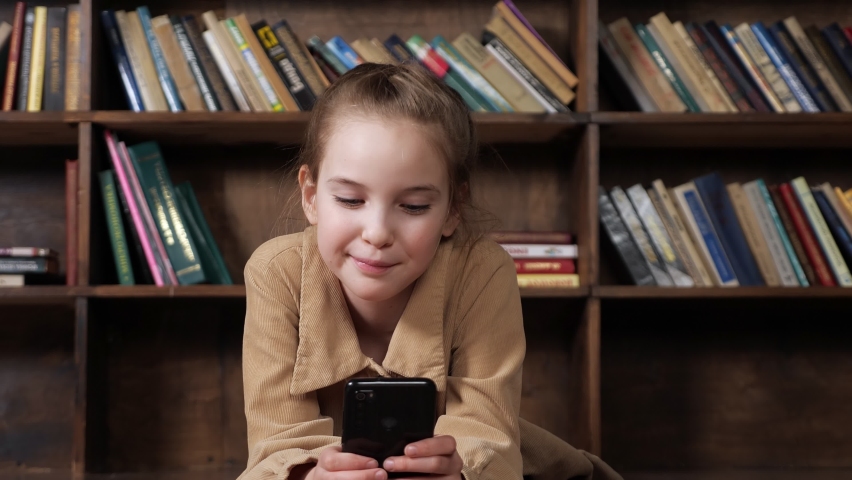 Girl junior student in brown jacket plays online game on black smartphone sitting against wooden bookshelves with coloured books in library