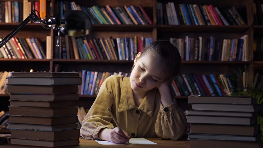 Tired girl writes home assignment on sheet of paper with pencil and yawns sitting among book stacks under electric lamp light in evening